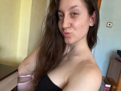 A young woman with long, dark brown hair and light brown eyes is posing for a selfie. She is wearing a black strapless top and has a small stud earring in her left ear. Her skin has some freckles and a mole on her cheek. Her lips are pursed. Her right arm is bent, and there are two red marks and a scratch on her elbow. Behind her, the wall is painted a light yellow on the left and a pale blue on the right. A wooden dresser with stacked clothes is visible on the right side of the image. There is also a small framed picture on the blue wall. A dark wooden desk is in the lower left corner. The overall setting appears to be an indoor space, possibly a bedroom. The location is identified as Popintsi, Bulgaria, although no specific landmarks or context are provided to confirm this.