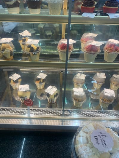 The image depicts a refrigerated retail display case in Lauro De Freitas, Brazil, showcasing pre-packaged desserts. The top shelf contains three multi-layered desserts in clear plastic cups and two chocolate cupcakes in red paper liners. The middle shelf presents five clear plastic cup desserts; two are topped with blueberries and light-colored fruit, and three feature red fruit and a granular base. The bottom shelf displays multiple clear plastic cup desserts: one with blueberries and light-colored fruit, two with blueberries, and several with dark speckled toppings. All individual dessert cups have white adhesive labels affixed to their lids. A red circular sign bearing the "Nestlé" brand logo is visible on the bottom shelf. In the lower right foreground, a large, transparent, circular container holds numerous small, white, powdered baked goods. This container has a white, scalloped-edge label with partially legible Portuguese text, including "Hummm que delícia!" and a "total" or "Ponto Total" logo. The display case features reflective glass shelves and metallic trim. The floor in the immediate foreground is dark and reflective, showing a distinct light reflection. No individuals are present in the scene.