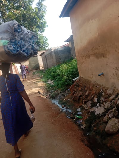The media file depicts a scene in Gwagwa, Nigeria. The primary subject is a woman walking away from the camera, carrying a large bundle of plastic water bottles on her head, secured in a dark mesh bag. She is wearing a vibrant blue and red patterned dress. To her left, another person, a man in a purple traditional outfit, walks further down the dirt path, followed by a child.

The setting appears to be a village or rural area, with a dirt road running alongside buildings constructed from mud and cinder blocks. Lush green vegetation grows on the right side of the path, where there is also a small, unmanaged drainage ditch filled with murky water and debris, including plastic bottles and wrappers. A blue pipe protrudes from the side of a mud-plastered wall. The sunlight suggests it is daytime, possibly late morning or early afternoon, with bright illumination and distinct shadows. The overall atmosphere is one of daily life and resourcefulness, with the woman actively transporting goods. No visible text is present in the image.