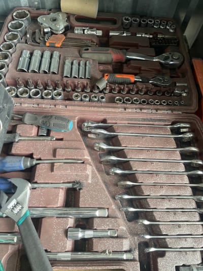 An overhead, full-frame, eye-level shot captures an open, dark red plastic toolbox filled with a variety of metal tools. The toolbox is positioned in a way that suggests it is on a flat surface. Dominating the frame are rows of sockets of various sizes, wrenches, ratchets, a screwdriver, and other miscellaneous tools. Some of the sockets are arranged in neat rows, while others are scattered. Wrenches are laid out horizontally in the lower right quadrant of the image, and ratchets are visible in the center of the frame. A blue and black handled screwdriver is positioned in the left half of the image. The tools appear to be made of silver-colored metal, with some having black or orange plastic handles. The surface of the toolbox itself is textured, with small white speckles visible throughout. The overall impression is that of a well-used, organized collection of tools. The location is identified as Сокуры, Russia.