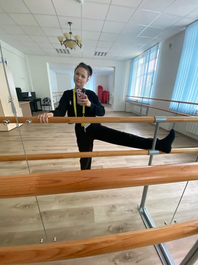 A young person, likely a teenager, is photographed from a low angle. The photo is taken indoors, with the young person positioned on wooden rungs, seemingly part of a ladder or some kind of gymnastic equipment. The person is wearing black pants, black socks, and a long-sleeved black shirt with yellow straps around the neck and shoulders, while holding a smartphone in their hand. The phone has a distinctive case. The background reveals an interior with a tiled white ceiling, a chandelier, and what appears to be office furniture. The location is in Peresvet, Russia, as indicated on the yellow lanyard.