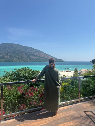 A full-height image depicts a single female figure standing on a balcony overlooking a tropical coastal scene in Ban Ko Li Pe, Thailand. The individual is viewed from the back, facing the ocean and a distant island. She has dark hair styled in a bun and is wearing a long, loose-fitting, olive-green garment with rolled-up sleeves. Her left hand rests on a dark-colored metal and glass panel railing. The balcony floor is composed of dark brown horizontal decking.
Below the railing, lush green tropical foliage is visible, including vibrant pink and red flowering plants on the left, and green bushes with yellow flowers on the right. Beyond the foliage, a white sand beach is present, featuring multiple beach umbrellas, lounge chairs, and several small, natural-material huts or bungalows. The ocean in the mid-ground exhibits clear turquoise shallow water transitioning into deeper blue. At least two traditional longtail boats are identifiable in the shallow water. A large, verdant, mountainous island or landmass occupies the left and central background. The sky above is clear and bright blue, without clouds. Shadows cast by the railing and the woman on the balcony floor indicate direct sunlight.