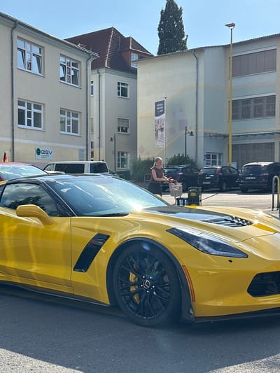 This image captures a bright, sunny day in Alsfeld, Germany. The main subject is a vibrant yellow sports car, likely a Chevrolet Corvette, parked on an asphalt road. Its sleek design and powerful presence dominate the foreground. Behind the car, a woman with blonde hair and glasses is visible, pushing a shopping cart. She appears to be casually dressed and is looking towards the car.

The scene is set against a backdrop of European-style buildings with cream-colored facades and numerous windows. Some windows are open, and one of the buildings features a large banner that reads "KITCHEN DAY" and another with the letters "SPD". The architecture suggests an urban or town setting. A tall tree with dense foliage is also present, adding a touch of nature to the urban landscape.

In the distance, other cars are parked in a row, including several dark-colored sedans and an SUV, indicating a public parking area or street. A yellow pole with a small flat object at the top extends upwards in front of one of the buildings. The overall atmosphere is relaxed and ordinary, suggesting a typical day. The bright sunlight casts distinct shadows, confirming it's daytime. The clear blue sky further reinforces the impression of good weather.
