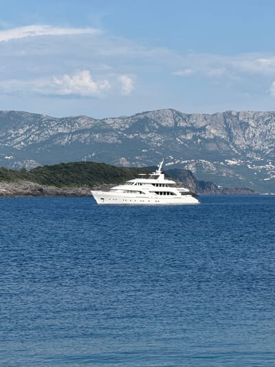This is a landscape photograph taken from a boat or elevated position, looking down at a coastline and the sea. The image is oriented vertically, with the sky at the left and the sea on the right.
The primary subjects are a large, white luxury yacht docked near a densely forested, rocky coastline. The yacht appears to be several stories high with multiple decks and windows. Behind the yacht and coastline, steep, rugged mountains rise, some with visible scattered buildings. The sea is a deep blue, with visible ripples indicating a calm or gentle breeze. The sky is a pale blue with scattered white clouds.
The scene depicts a tranquil moment with the yacht anchored in clear, blue waters, likely a popular spot for leisure and tourism. The time of day appears to be daytime, possibly late morning or early afternoon, given the brightness and clear sky. The weather is sunny and pleasant. The location cues (Dubraljevina, Montenegro) suggest a coastal area known for its natural beauty, combining mountainous terrain with the Adriatic Sea.
There is no visible text in the image. The overall mood is one of serenity and luxury, showcasing the juxtaposition of natural grandeur and man-made opulence.