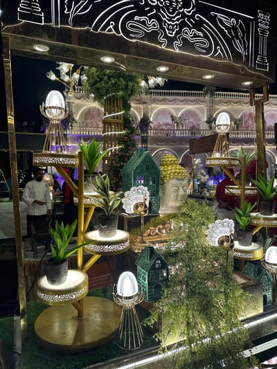 Intricate decorations adorn an ornate space. A golden Buddha head sits amid lush greenery, decorative houses, and floral arrangements. Unique lighting fixtures add ambiance with pineapple plants. An elaborate wall panel with glowing designs stands adjacent to architectural features. A chef and dining staff are visible in the background, hinting at a restaurant setting.