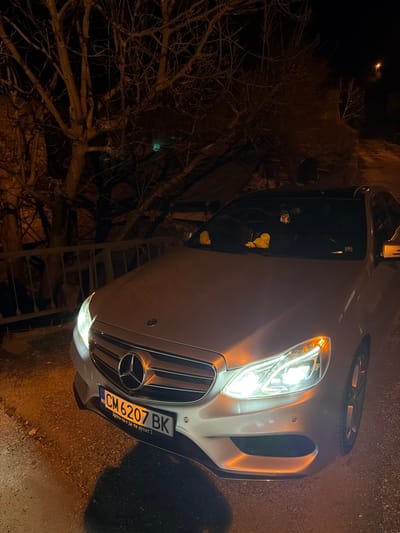 The image captures a silver-colored Mercedes-Benz sedan parked at night in Podvis, Bulgaria. The vehicle is angled towards the viewer, with its headlights illuminated, projecting bright white and amber light. The front bumper displays a Bulgarian license plate, "CM 6207 BK." On the dashboard, visible through the windshield, are two yellow, duck-shaped figures and a small, dark object suspended near the rearview mirror. The car features a dark-colored roof.
In the background, to the left of the vehicle, stands a large, leafless deciduous tree with a dense network of bare branches. A light-colored metal fence or railing runs horizontally between the car and the tree line. The ground beneath the car is dark and appears to be an uneven, possibly paved or gravel, surface. In the upper right background, a distant street light emits a faint orange glow. A subtle, diffuse green light source is barely discernible through the tree branches on the left side of the frame. No individuals are present.