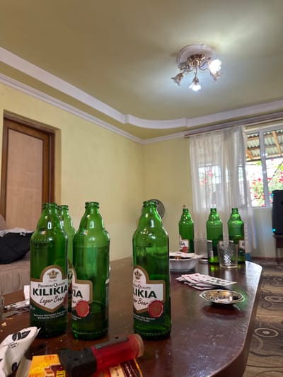 The image depicts a table laden with several empty green bottles of Kilikia Lager Beer, a brand known to have been produced in Aygevan, Armenia. The bottles are lined up in a row, some with their labels clearly visible. The setting appears to be indoors, possibly in a living room or dining area, indicated by the presence of a light fixture on the ceiling, a window with curtains, and a glimpse of interior furnishings. On the table, besides the bottles, there are playing cards, an ashtray with cigarette butts, and other miscellaneous items.