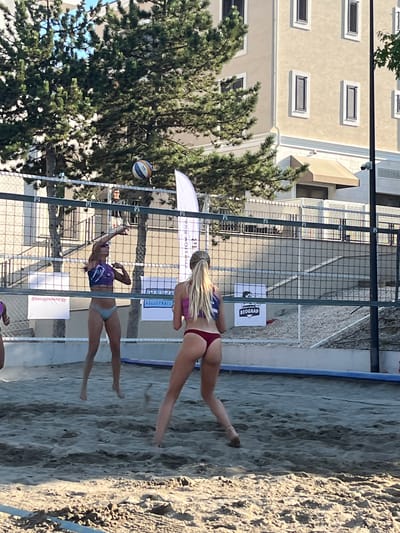 Two women are playing beach volleyball on a sand court in Gošići, Montenegro. The woman on the left is wearing a purple and blue bikini top and a light blue bikini bottom. She is in the process of serving the ball. The woman on the right is wearing a pink bikini top and a maroon bikini bottom. She is facing away from the camera and appears to be preparing to receive the serve. A volleyball is in the air above the net. The court is bordered by a fence and a building with light-colored walls and dark shutters can be seen in the background. Trees with green foliage are also present. The scene is outdoors and appears to be during daylight hours.