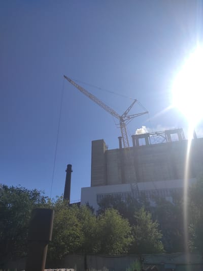 The image shows a large industrial facility with a tall chimney and a construction crane against a clear blue sky. A bright sun is visible in the upper right corner, causing lens flare. The building appears to be made of concrete and has large windows. There is some steam or smoke rising from the top of the building. The foreground shows trees and a dark, cylindrical structure. The overall scene suggests an active or recently active industrial site, possibly undergoing construction or maintenance. The specific location in the city of Kyzyl, Kazakhstan, is not discernible from the image itself, as it lacks any identifying landmarks or textual information.