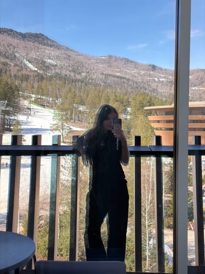 A reflection in a window captures a woman holding a phone while standing on a balcony with a view of mountains and a ski resort. The reflection includes part of the balcony's structure, featuring horizontal slats. The backdrop is dominated by a mountain covered with trees and patches of snow, under a clear blue sky. Ski slopes are visible below the mountain. The exact location in Озёрное, Russia cannot be determined from the image.