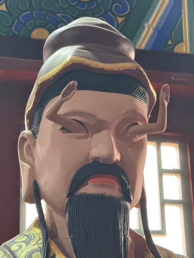 The image shows a close-up of a statue's head. The statue has light skin, a black mustache, and a black hat with gold trim. Two snake heads emerge from the head near the eyes. The background includes a window and a decorative painted ceiling with green, blue, and gold patterns. It's impossible to verify the statue's location in Lintong District, China, based solely on the image.