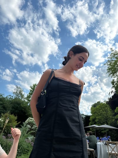 The image shows a woman with dark hair pulled back, wearing a black dress and a small black purse, positioned at an angle against a backdrop of a partly cloudy sky. Her face is turned towards the upper right of the frame, and she appears to be speaking or smiling. In the background, there's a glimpse of a garden with various plants and flowers. To the right, there are hints of a table set for a meal with blue floral arrangements, and people seated. A hand is visible in the bottom right corner. The location, based on the presence of manicured gardens, a setting for outdoor dining, and the architectural style of any unseen buildings, could possibly be somewhere in the city of Cambridge, United Kingdom.