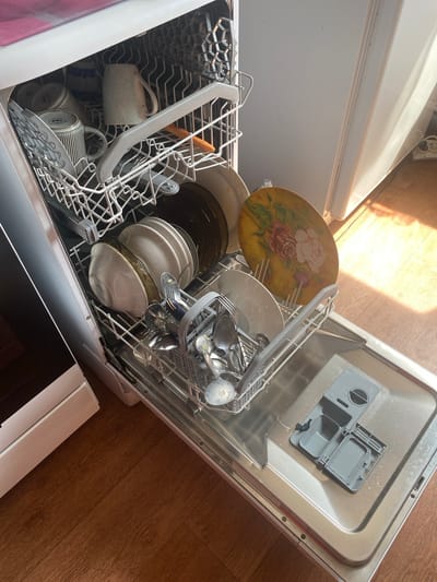 The image shows an open dishwasher filled with dishes and silverware. The dishwasher is white and appears to be a standard household appliance. It is located on a wooden floor, which is partially illuminated by sunlight coming from the right side of the frame. The dishwasher contains several items, including mugs, plates, bowls, and cutlery. One of the mugs has a striped pattern, and another is plain white. There are also several plates stacked in the upper rack. The lower rack contains a cutlery basket filled with spoons, forks, and knives. A decorative plate with a floral pattern is also visible in the lower rack. The detergent dispenser is located on the front of the dishwasher door. The overall scene depicts a typical domestic setting, likely a kitchen. The location is Urazovo, Russia.