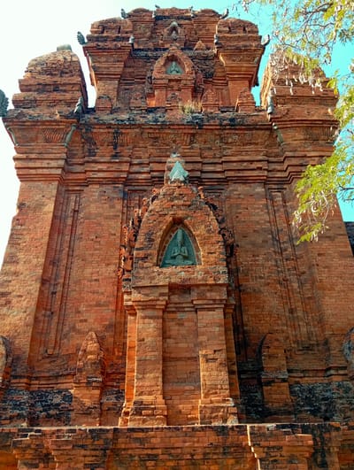 The image captures a majestic, ancient brick tower, likely a Cham temple, viewed from a low angle looking upwards. The structure is predominantly made of reddish-brown brick, exhibiting signs of age with darkened patches and some vegetation growing in crevices. Its design is intricate, featuring multiple tiered levels, decorative corbelling, and recessed panels.
Several carved figures are integrated into the brickwork. A prominent one is centered towards the middle of the tower, seated within an arched niche, depicted in a meditative pose. Another similar figure is visible higher up, suggesting a repetitive architectural motif. The tower also features smaller decorative elements and spires at its peak.
The setting is outdoors under a clear, bright blue sky, visible on the right side and upper background. Bright sunlight illuminates the bricks, casting strong shadows that highlight the texture and three-dimensional qualities of the architecture, suggesting a sunny day, possibly morning or late afternoon. Green foliage from a tree is partially visible on the right side, with some branches extending into the frame, adding a natural element to the scene. There is no discernible human activity or visible text in the image. The structure's grandeur and weathered appearance evoke a sense of history and cultural significance, characteristic of the Cham towers found in Phan Rang – Tháp Chàm, Vietnam.