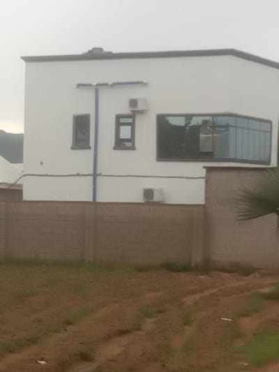 The image shows a modern white building with a distinctive angular design. The building has multiple windows, including a large, angled window on the upper floor that reflects the sky and some greenery. There are also smaller rectangular windows on the lower floors. Two air conditioning units are visible mounted on the exterior wall, one above the other. Blue pipes run vertically and horizontally along the white facade, suggesting some kind of utility or plumbing system.

To the right of the building, a textured brick wall is visible, and beyond that, an area of uneven, reddish-brown ground covered with sparse green vegetation. This ground appears to be an undeveloped or partially developed plot of land. A palm frond is partially visible in the upper right corner of the image, suggesting a tropical or subtropical climate.

The overall lighting suggests it might be late afternoon or early morning, with the sky appearing overcast or hazy. There are no people or obvious signs of activity in the image. The scene depicts a residential or commercial building in a somewhat rural or undeveloped setting, possibly in Zanzi, Nigeria, given the location context. The image is oriented vertically, with the top of the building appearing on the left side.
