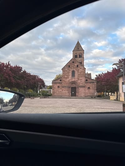 The image displays the Église Sainte-Croix, located on Place de l'Hôtel de Ville in Kaysersberg-Vignoble, France. The perspective is from inside a vehicle, indicated by the dark gray dashboard visible in the lower foreground and the car's interior frame arching across the top. The main subject is the Romanesque church, constructed from reddish-brown stone, featuring a prominent central bell tower with a pointed spire capped by a cross. The church's facade includes a large arched main entrance door and multiple arched windows. It stands on a wide, empty, paved stone plaza. To the left of the plaza, a row of deciduous trees with dark red and brown autumn foliage is visible, alongside several rectangular planters. A side-view mirror on the left reflects a street with parked cars and more distant buildings. To the right of the church, a streetlamp and part of another building are visible amidst similar autumn foliage. The sky is overcast with scattered clouds. There are no individuals present in the plaza or directly surrounding the church.