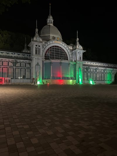The image depicts the Zheleznovodsk Spa House, a historical building located in Zheleznovodsk, Russia. The facade of the building is illuminated by green and red lights, creating a striking contrast against the night sky. The building features ornate architectural details, including a central dome, arched windows, and intricate metalwork. The foreground is a paved courtyard, also dimly lit. The overall scene suggests a nighttime view of a significant cultural landmark.