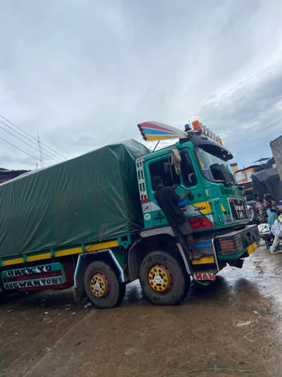 In Ogbunike, Nigeria, a man is seen climbing the side of a green truck that has a large green tarp covering its cargo. The truck has bright yellow, red, and blue accents on its body and wheels. A colorful, wing-like decoration with red, blue, and yellow stripes is attached to the top of the truck's cab, along with several flashing lights. The truck has "Unoguway Transporter" written on the side of the cab. The ground is wet and appears to be made of dirt or mud, with scattered debris. In the background, other trucks and people are visible, suggesting a busy marketplace or loading area. The sky is overcast.