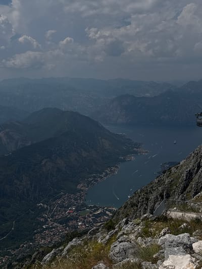 This is a vertically oriented, high-angle photograph overlooking a mountainous landscape and a body of water. The scene appears to be a coastal or lakeside region with steep, rugged cliffs and a town nestled along the shoreline. The terrain is densely covered with vegetation, primarily trees, giving the mountains a dark, verdant appearance.
In the water, several boats are visible, leaving white wakes, suggesting they are moving. The town, characterized by its reddish rooftops and dense architecture, is spread across the lower slopes of the mountains, descending towards the water. One notable structure with a bright red roof stands out amongst the other buildings.
In the foreground on the right, there are large, grey rocks, some with sparse green vegetation growing between them. A narrow path or walkway, bordered by a metal railing, is visible built into the cliff face, suggesting a scenic viewpoint or hiking trail.
The sky is partly cloudy, with large, fluffy white clouds against a pale blue backdrop. The lighting suggests it is daytime, likely mid-morning or afternoon, with the sun casting shadows that highlight the topography of the mountains. The overall mood is one of natural beauty and tranquility, with a hint of human settlement integrated into the rugged environment. There is no visible text in the image.