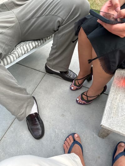 The image shows a partial view of three people seated outdoors on a gray tiled surface. One person wears gray pants and black penny loafers with white socks, the second wears a black dress and strappy heeled sandals with purple, green, and pink accents, and the third wears white pants and blue flip-flops. A small gray table is partially visible in the upper center of the image. The specific location in Cambridge, United Kingdom, cannot be determined based on the available visual information.