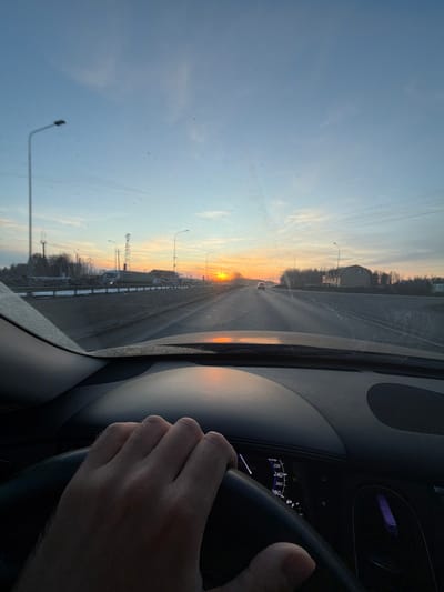 The image presents a first-person view from the interior of a vehicle traveling on a multi-lane road in Beloborodovo, Russia, during what appears to be sunrise or sunset. The sky transitions from a light blue overhead to a prominent orange and yellow gradient at the horizon, with the sun visible as a distinct orange orb on the right side of the frame. The vehicle's windshield exhibits numerous dust or dirt particles and some streaking.

In the lower-left foreground, a human right hand, displaying light hair on the wrist, grips the dark, possibly leather, steering wheel. The dark dashboard occupies the lower portion of the image. Through the steering wheel, a segment of the vehicle's instrument cluster is visible, displaying speed increments from 200 to 280 km/h and a fuel gauge icon.

The road ahead is a dark, multi-lane surface, possibly damp, with white lane markings receding into the distance. On the left side of the road, beyond a gray guardrail with patches of snow or ice, are bare trees, multiple utility poles, and at least two communication or industrial towers. A large vehicle, possibly a truck, is partially visible behind the guardrail. Several streetlights line the left side of the road. On the right side of the road, also beyond a guardrail, stands a line of bare trees, another utility pole, and a light-colored, multi-story building. A white sedan is observed traveling ahead in the same lane in the mid-distance.