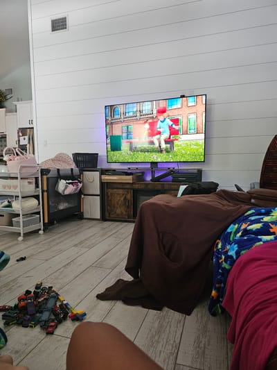 The image depicts an indoor scene, likely a living room, with a large television screen displaying a cartoon. The room is furnished with a TV stand, a playpen, storage shelves, and a pile of toy cars on the light-colored wooden floor. Two blankets, one brown and one with a dinosaur pattern, are draped over a piece of furniture or a person. A portion of a person's leg is visible in the foreground. The walls are paneled with white horizontal planks. A vent is visible high on the wall. The overall impression is a casual, family-oriented living space. The city of Islamorada, United States of America is mentioned, but there are no specific visual cues in the image to confirm or deny this location.