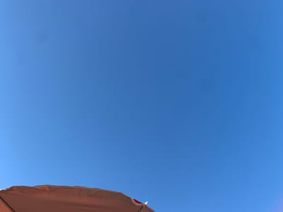 This is a close-up, low-angle shot of a portion of a peach-colored umbrella against a clear, bright blue sky. The umbrella is positioned in the bottom portion of the frame, its edge creating a jagged, organic line across the lower third of the image. The fabric of the umbrella appears to be slightly wrinkled and stretched, suggesting it's open and taut. There are faint shadows and highlights on the umbrella's surface, indicating direct sunlight.

The sky is a uniform, vibrant blue with no clouds, suggesting a clear and sunny day. There are two faint circular smudges or lens flares visible in the upper left quadrant of the sky, which are likely artifacts from the camera or lens.

The overall impression is one of a bright, summery day, possibly at a beach or outdoor gathering, given the presence of the umbrella. The low angle and focus on the umbrella against the expansive sky create a sense of openness and perhaps a feeling of being sheltered from the sun. There is no visible text in the image. The time of day appears to be daytime, likely mid-morning to late afternoon, due to the intensity of the sunlight. The location context of Badalona, Spain, further supports the idea of a warm, Mediterranean climate.