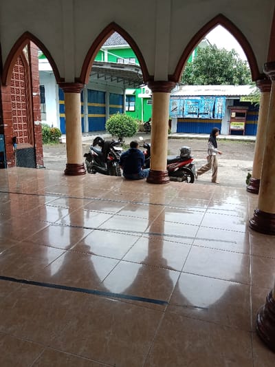 The image shows a tiled courtyard with a shiny surface reflecting the surrounding environment. Two men are sitting on the ground, with one man in the middle of the frame, facing left, and the other man further back and slightly to the right. In the background, a woman in a hijab is walking away from the camera on a paved path to the right of the image. To the left of the courtyard is a black motorcycle with a red seat. To the right, another motorcycle with a red seat faces toward the camera. The courtyard is surrounded by columns and arches, creating a shaded area. Behind the arches is a small building with blue and green painted walls. Green plants are growing in the area. The coordinates (latitude: -6.948, longitude: 110.231) indicate the scene is located in the Kecamatan Brangsong area of Indonesia.