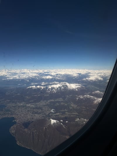 The image presents an expansive aerial view captured from an airplane window, showcasing a dramatic landscape of mountains, a body of water, and an urban settlement under a clear blue sky.

The upper half of the frame is dominated by a vast, clear, and deep blue sky, indicating bright, sunny weather conditions at high altitude. Below the sky, stretching across the middle of the image, is an extensive range of snow-capped mountains. These mountains feature numerous jagged peaks and ridges, all covered in brilliant white snow, with darker rock faces visible beneath the snow in many areas. Scattered white clouds are interspersed among the mountain peaks, some partially obscuring distant ranges, adding depth to the scene.

Towards the lower left quadrant of the image, a large body of dark blue water, resembling a lake or a wide bay, is visible. Along its shores and extending inland, a densely populated urban area or city sprawls. The city appears as a mosaic of buildings and streets, blending into the surrounding terrain. Between the city and the main snow-capped mountain range, there are lower, darker hills and mountains, some showing patches of snow, suggesting a transition from urban areas to higher elevations.

The dark, curved frame of an airplane window is visible along the right edge and bottom right corner of the image, partially framing the view and indicating the perspective. Minor smudges or dust specks are discernible on the window surface, particularly noticeable against the clear sky in some sections, which are characteristic of photographs taken from an aircraft. The overall impression is one of vastness and natural grandeur seen from a unique vantage point.