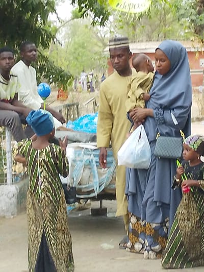 The image depicts a family and other individuals outdoors, possibly at a market or public space in Garun Gawa, Nigeria. Two young men are seated on a structure adorned with a blue balloon. A man in a yellow robe and a woman in a blue hijab are standing. The man is holding a young child. The woman is carrying a white plastic bag and a purse. Two young girls in matching patterned dresses stand to their right. The background contains trees and indistinct figures.