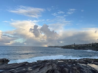 A wide-angle, eye-level shot captures a coastal landscape with a view of the ocean and a town nestled on a cliffside. The foreground is dominated by dark, textured rocks, some of which are wet and reflective, suggesting recent rain. In the mid-ground, the deep blue ocean churns with white-capped waves breaking against the rocky shore. A faint rainbow arcs across the water in the distance, a subtle indication of recent or ongoing precipitation. The horizon line is clearly defined, with a cluster of buildings forming the town on the distant cliff. The sky above is a mix of bright blue patches and dramatic, billowing clouds, some tinged with yellow and gray. The lighting suggests it is either early morning or late afternoon. The scene appears to be a natural coastal setting in Clovelly, Australia, with the town of Clovelly visible on the cliffs. There are no people or animals visible in the frame.