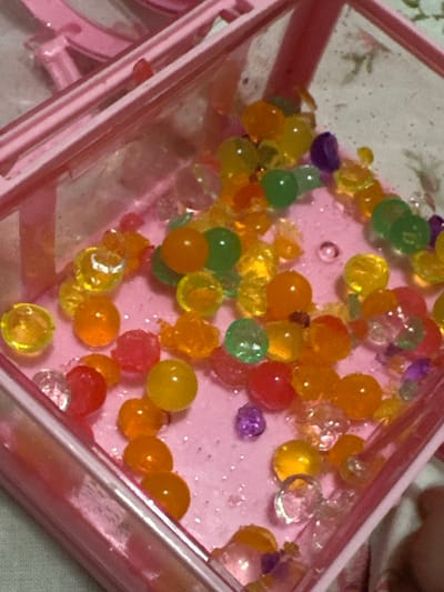 A close-up shot of a pink plastic container filled with an assortment of colorful water beads of various sizes. The beads are primarily translucent shades of green, yellow, orange, red, and clear, scattered randomly across a light pink base. The pink container is angled slightly, showing the edge and corner details of its structure. There is an absence of people in the scene, and no actions or interactions are taking place. The environment is limited to the contents of the container and the immediate surrounding area, which appears to be a flat surface. The image does not provide any geographic context related to Uva, Russia.