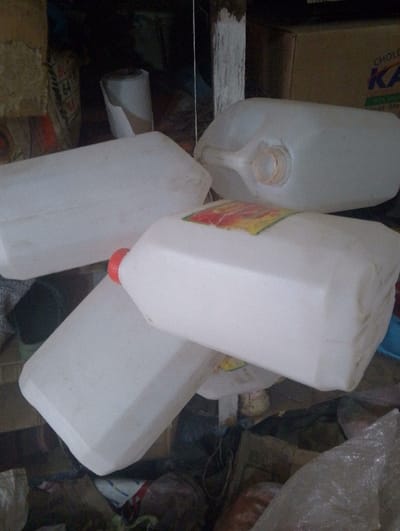 The image depicts a cluttered indoor storage area within Kinale ward, Kenya. Multiple white, rectangular plastic jerrycans or containers are prominently displayed, stacked or positioned haphazardly. One jerrycan in the upper right is lying on its side with its cap removed, revealing a circular opening and an integrated handle. Another jerrycan in the center foreground features a red cap and a partially visible yellow and red label. The background is dimly lit and contains various indistinct objects, including cardboard boxes; one box displays partial text "CHOLE..." and "KA". A white-painted wooden post or beam is visible vertically on the right side. No individuals are present, and no actions or interactions are occurring.
