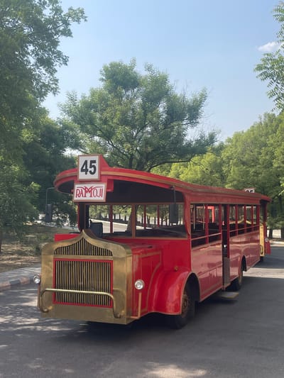 A double-decker red bus with gold-colored detailing is parked in an outdoor location. Signage on the bus includes the number "45" and the word "RAMOJI." The bus is surrounded by trees and greenery. The location is most likely Ramoji Film City in Anajpur, India. The bus appears to be a stationary prop or display rather than in active use for transportation. The surrounding area is a paved road or walkway.