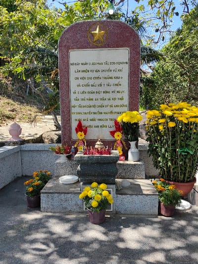 The image shows a memorial with a red stone stele marked with a gold star. The plaque commemorates the events of March 1, 1968, where vessel 235 of HONDVN carried out a weapons transport mission to support battlefront area 6. It battled 7 warships and 2 American-backed allied special forces units, managed to safely deliver the cargo, and scuttled the ship. Fourteen officers and soldiers died heroically, including Captain Nguyen Phan Vi. The memorial also states that the ship was posthumously awarded the title of Heroic Unit by the state. Flowers and offerings adorn the site.