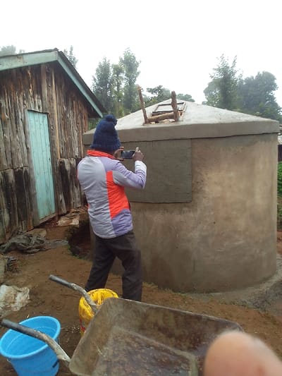 The image depicts an outdoor scene in Ihururu, Kenya, centered on an individual documenting a concrete water storage tank. A single person, viewed from the back, stands slightly left of center, wearing a dark blue knit beanie, a multi-paneled jacket with grey, purple, and orange sections, and dark trousers. This individual holds a black smartphone horizontally with both hands, aiming it towards the large, cylindrical concrete water tank on the right.

The concrete tank features a conical top with two unfinished wooden posts extending vertically from an opening. A rectangular area of lighter concrete, possibly wet or a patch, is visible on its side. To the left of the person, a rustic building with vertical wooden plank siding and a light blue wooden door is present.

In the immediate foreground, a rusty wheelbarrow with wooden handles rests on the uneven dirt ground. To its left, a yellow plastic bucket and a light blue plastic bucket are also situated on the ground. A human finger is partially visible in the bottom right corner of the frame. The background consists of dense green foliage under an overcast sky, indicating an outdoor, possibly rural or semi-urban environment.