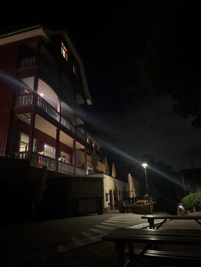 The image captures a nighttime outdoor scene in what is identified as Heidelberg, Germany, specifically a Berggasthof (mountain guesthouse) or similar hotel establishment, indicated by the visible "BERG-G... HOTEL" signage.

The primary subject is a multi-story building positioned on the left, featuring a dark red wooden facade with white architectural trim and white-railed balconies spanning at least three visible levels. Several windows on the building's upper and lower floors emit a warm, yellow light. To the right, the building transitions to a section with a lighter, possibly cream-colored facade, where multiple large, beige-colored outdoor umbrellas are neatly folded.

In the midground, a paved pathway or street runs horizontally, with a section marked by illuminated white stripes resembling a crosswalk. A tall, black street lamp stands on the right side of this path, emitting a bright, warm light that creates a distinct lens flare diagonally across the frame towards the upper left.

The immediate foreground on the right side features two dark wooden picnic tables with integrated benches. Further along the pathway, adjacent to the lighter-colored building, additional outdoor tables and chairs are visible, accompanied by a large wooden barrel. The background on the right is dominated by dark, dense foliage and trees, silhouetted against a dark night sky with discernible cloud cover and faint celestial points. No individuals are present in the scene.