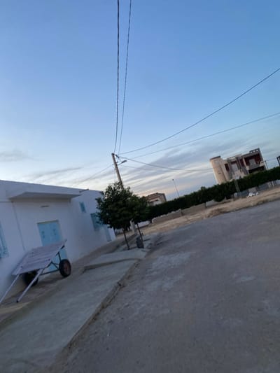 A street scene under a blue sky. A white building with blue-framed windows is on the right, with a wheelbarrow leaning against the wall. Further down the street are trees and taller buildings. Power lines stretch across the sky from a pole near the white building. The road surface is concrete.