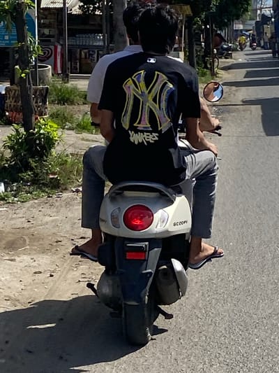 The image shows two people on a scooter driving down a street in Tingkeummanyang, Indonesia. The person in the back is wearing a black t-shirt with a large graphic of the New York Yankees logo. They are also wearing grey pants and flip-flops. The person in the front is wearing a white shirt. The scooter is white and has a red taillight. The street is paved and there are buildings on either side. There are also trees and vegetation visible in the background. The sky is blue and there are some clouds. The image appears to be taken during the day.