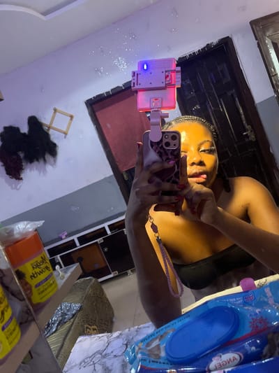 The image displays an indoor mirror selfie taken in Gbongan, Nigeria. A young woman, visible from the chest up, is reflected in a mirror. She has dark braided hair, wears makeup, and is dressed in a dark strapless top. Her right hand, with red manicured fingernails, holds an iPhone-like smartphone in a purple and pink leopard print case. A pink, clip-on ring light with a glowing blue indicator is attached to the top of the phone, and a pink braided wrist strap hangs from the device. The woman's head is slightly tilted, and her tongue is partially extended.
The foreground surface, exhibiting a white marble-like pattern, holds several items. On the left, an orange bottle with a yellow label, partially readable as "Mega Growth," is present. To the right, a blue package of "Angel" brand wet wipes with a blue flip-top lid is visible.
The reflected background reveals a room with light-colored walls, appearing pinkish-purple due to lighting. On the left wall, several dark hair extensions hang, alongside a light-colored wooden drying rack. Below these, a dark wooden cabinet or dresser with multiple compartments is situated. A dark wooden door with a metallic handle is reflected on the right side. The ceiling is white with decorative molding, and scattered bright points of light or glitter are visible on the upper reflected wall.
