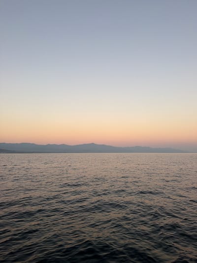 The image depicts a serene seascape at dusk, with the sky transitioning from a pale blue at the top to soft hues of orange and pink near the horizon. A range of hazy, mountainous terrain is visible in the distance, separating the sky from the water. The foreground is dominated by the dark, rippling surface of the ocean, reflecting the muted colors of the sky. The scene is devoid of any people, structures, or discernible activity, creating a sense of natural tranquility. The overall impression is one of a quiet, expansive natural landscape.