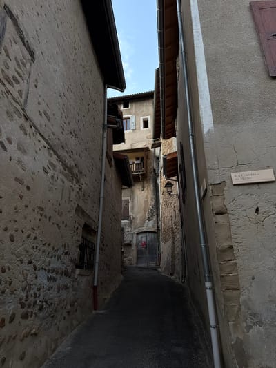 The image shows a narrow cobblestone alleyway in Saint-Antoine-l'Abbaye, France. The alley is flanked by old stone buildings with weathered facades. A street sign on the right building reads "Rue Corsiere ou du Milieu." A dark wooden door is visible at the end of the alley. The sky is visible at the top of the frame, with a few clouds.