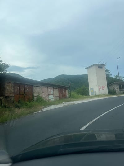 This is a photograph taken from a vehicle, likely in the city of Pechinska, Bulgaria. The scene depicts a rural or semi-rural roadside environment with a cloudy sky overhead.

In the foreground, a dark asphalt road with a white dashed line is visible, indicating a traffic lane. The dashboard of the vehicle from which the photo was taken is partially visible at the bottom of the frame, reflecting some of the surroundings.

To the left of the road, a long, dilapidated brick building with several large, rusty metal garage doors is situated. Overgrown grass and weeds are present in front of and around this building. The structure appears old and weathered.

Further down the road, to the right, stands a tall, rectangular, light-colored concrete structure, which appears to be a transformer station or an electrical substation. It has a small opening near the top and electrical conduits or wires running to and from it, connecting to overhead power lines. Red graffiti, reading "P 21," is present on the front of this structure. Adjacent to the transformer station is a smaller, windowed brick building, also showing signs of age.

The background of the image is dominated by lush, green, rolling hills covered in trees, suggesting a mountainous or hilly terrain. The overall impression is one of a somewhat isolated, utilitarian roadside area set against a natural landscape. There are no people or animals visible in the scene.