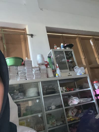 The image shows an indoor scene of a room that appears to be a small shop or storage area. The room is filled with shelves stacked with various items. In the foreground, a person wearing a black hijab is partially visible on the left side of the frame.

On the shelves, there are stacks of cigarette packs, green plastic basins, rolls of white material (possibly plastic wrap or foam), and bottles of cleaning supplies. There is also a glass-fronted cabinet on one of the shelves containing more items, some of which are indistinct. The background consists of wooden partitions or walls, suggesting a simple construction. The lighting appears to be artificial and somewhat dim, casting shadows. The overall impression is one of a cluttered but functional space, possibly a local convenience store or a personal storage area in Unnyi, Indonesia.
