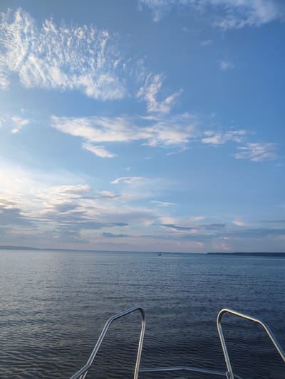 The image depicts a calm body of water, likely a lake or large river, under a partly cloudy blue sky. The perspective is from the front of a boat, with two polished metal handrails visible in the foreground. The water's surface is gently rippled, reflecting the sky. In the distance, a tree-lined shore is visible under a band of clouds tinged with the soft light of either sunrise or sunset. A small boat is present on the water in the mid-ground, to the left of the frame. The overall impression is one of peaceful tranquility. No people are directly visible in the image. The location is stated to be the city of Galevo, Russia.