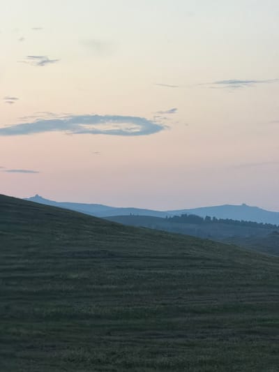 The image depicts a serene, open landscape at dusk, likely in the vicinity of Кусеево, Russia. The sky is a soft gradient of pale yellow and pink hues, with a few wispy clouds scattered across it. Below the sky, a series of rolling hills are visible, fading into a hazy blue in the distance. The foreground is dominated by a grassy hillside, rendered in dark green tones, suggesting it is either late evening or early morning. The texture of the grass indicates a natural, possibly untamed, terrain. The overall atmosphere is peaceful and tranquil, with no signs of human presence or activity.