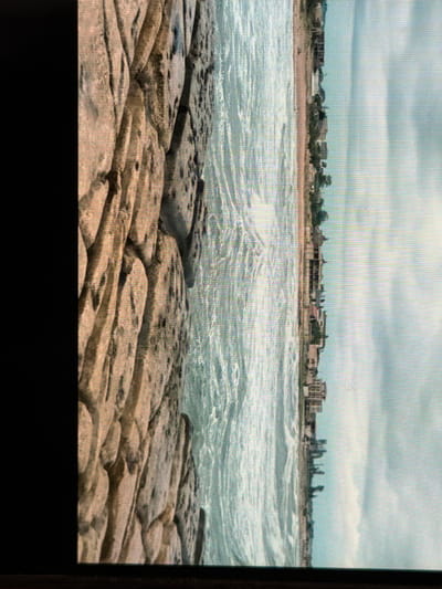 The media file depicts a coastal scene with a rocky foreground, the sea, and a distant shoreline with buildings under a cloudy sky. The foreground is dominated by large, flat, irregularly shaped rocks, some of which appear wet, suggesting they are near the water's edge. The sea is choppy with visible waves and white foam, indicating rough conditions or a strong current. In the background, a sandy beach separates the sea from a line of buildings. These buildings appear to be a mix of residential and perhaps some commercial structures, with varying architectural styles. Trees are scattered along the shoreline and behind the buildings. The sky is overcast with thick, gray clouds, suggesting it is either a dull day or nearing sunset. The overall impression is one of a natural landscape with human habitation. There is no visible text in the image. The scene does not appear to be actively populated by people, though a very small, indistinct figure might be present on the beach in the middle distance.