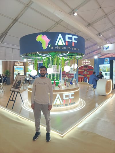 This media file captures a man posing in front of a modern exhibition booth at an indoor event, likely a trade show, in Al Machouar – Stinia, Morocco. The man, standing in the foreground, wears a grey sweatshirt with "Calvin Klein" emblazoned on it, grey sweatpants, and black sneakers. He has a beard and looks directly at the viewer.
The central exhibition booth is circular, featuring a large overhead sign that prominently displays "AFC a vision to share" alongside a vibrant, stylized map of Africa. The booth is decorated with hanging artificial greenery and features round white pendant lights. A smaller, illuminated circular sign at the base of the booth also reads "ACC vision to share". QR codes are visible on the booth's decorative elements, suggesting modern information access.
In the background, other attendees are visible, some seated at tables and counters, engaging in discussions or observing the exhibits. To the left, a framed portrait, likely of a Moroccan dignitary, is displayed on an easel. Adjacent booths feature branding such as "Jibal", "Kayna" (showcasing packaged products), and "B BAG-IN" with the Arabic text "تأمين ، حماية ، تن ما" (meaning "Assurance, protection, growth"). The venue has a bright, professional atmosphere, characterized by a white fabric tent ceiling, exposed metal trusses, and cylindrical air ducts. The booth itself features contemporary design elements, including integrated strip lighting along its base and floor edges.