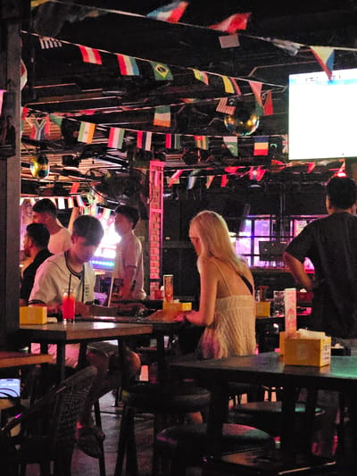 The image shows a group of people in what appears to be an indoor establishment, possibly a bar or restaurant, with a dark, atmospheric setting. Numerous small flags of various countries are hanging from the ceiling and walls, creating a festive and international ambiance. The lighting is dim with colorful neon lights, suggesting it's nighttime.

In the foreground, several individuals are visible. On the right, a woman with blonde hair is leaning forward, wearing a white top. To her left, a person in a white t-shirt is visible, looking down. Below them, another person in a white t-shirt with dark trim is also looking down, with a third person in the immediate foreground, also looking downwards. The people seem engaged in conversation or watching something.

In the background, more people are dimly lit. There are also shelves with various items, including what look like bottles and boxes, with some illuminated by colorful lights. A large screen is visible at the top left, displaying bright white content. The overall scene suggests a lively gathering, perhaps a tourist spot given the international flags. The expressions on the faces of those clearly visible are not discernible due to the lighting and angle. The environment is indoors, and the time of day is implied to be evening or night due to the artificial lighting and dark surroundings. There are no clear indications of weather. No specific text is legible within the image, except for what appears to be a logo or characters on a yellow box in the mid-right background. The location context provided is Ban Klong Haeng, Thailand.