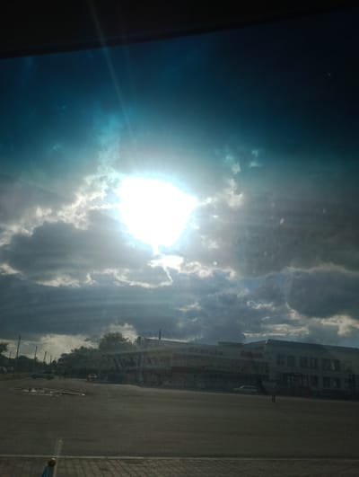 The image captures a cloudy sky with the sun shining brightly through a gap in the clouds, casting rays of light downwards. Below the sky, a street scene is visible, featuring a building with multiple storefronts, including "Sinsay" and "Shikpak." Several cars are parked or driving on the street and in front of the building. Trees line the street in the background. The foreground shows a paved area, possibly a parking lot or a pedestrian walkway, with some visible puddles of water. The overall impression is of an outdoor commercial area in Atbasar, Kazakhstan, under a dramatic sky.
